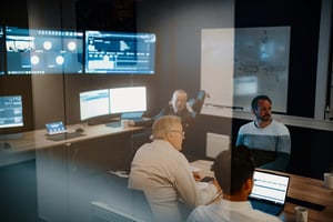 Group of engineers in a control room reviewing real-time data on multiple monitors. Some are engaged in discussion while others observe. Reflections in the glass show additional screens and digital interfaces